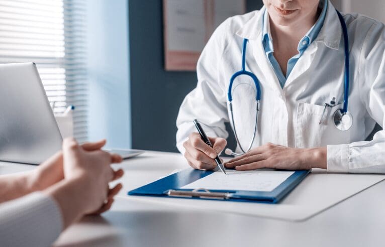 A doctor wearing a white coat and stethoscope writes on a clipboard during a consultation. A patient sits across the table, hands clasped. A laptop is open nearby.